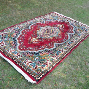 May include: A red, green and gold patterned rug with a white border. The rug has a floral design and is laid out on a green grassy surface.