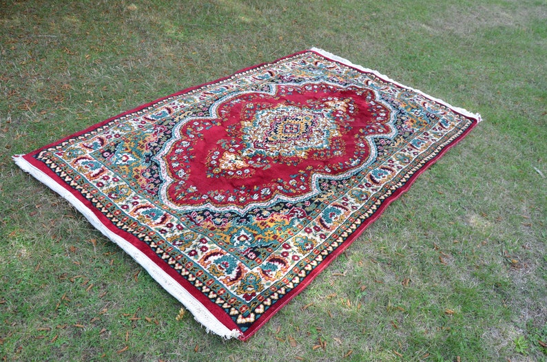 May include: A red, green and gold patterned rug with a white border. The rug has a floral design and is laid out on a green grassy surface.