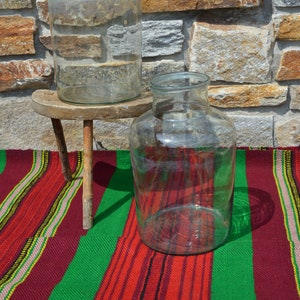 May include: Two clear glass jars of different sizes. One jar is on a small, rustic wooden stool. The other jar sits on a colorful striped textile with red, green, and yellow stripes. The background is a stone wall.