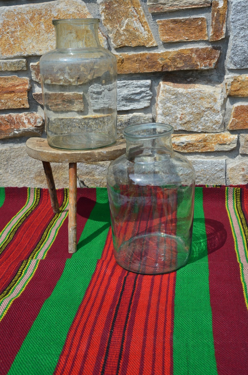 May include: Two clear glass jars of different sizes. One jar is on a small, rustic wooden stool. The other jar sits on a colorful striped textile with red, green, and yellow stripes. The background is a stone wall.