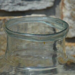 May include: A clear glass jar with a wide, rounded base and a narrow neck. The rim of the jar is smooth and slightly curved. The jar is made of transparent glass, allowing a view of the contents. The background is a stone wall.