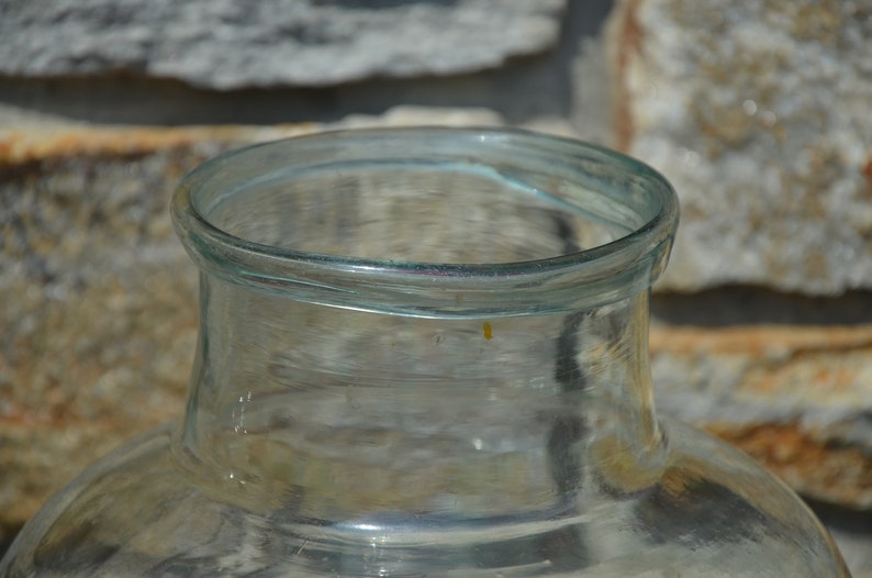 May include: A clear glass jar with a wide, rounded base and a narrow neck. The rim of the jar is smooth and slightly curved. The jar is made of transparent glass, allowing a view of the contents. The background is a stone wall.