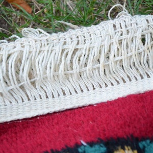 May include: Close-up of a white fringe on a red rug with a colourful pattern. The fringe is woven with a simple knot pattern.