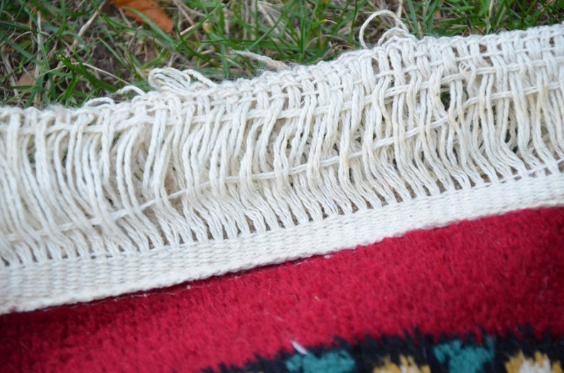 May include: Close-up of a white fringe on a red rug with a colourful pattern. The fringe is woven with a simple knot pattern.