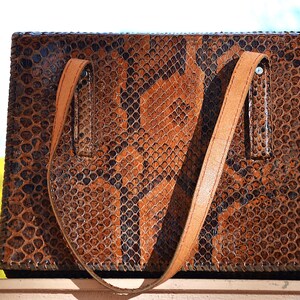 Könnte beinhalten: Rechteckige Handtasche mit Schlangenmuster in Braun- und Schwarztönen. Die Tasche hat zwei hellbraune Griffe und ein strukturiertes Design, das auf einen Vintage- oder klassischen Stil hindeutet. Die Tasche steht auf einer Holzoberfläche.