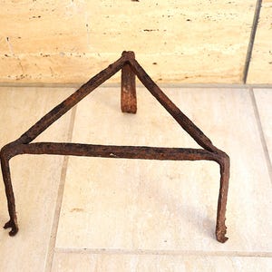 Può includere: Un sottopentola triangolare in metallo arrugginito con tre gambe. Il sottopentola è realizzato in metallo marrone scuro e ha un aspetto invecchiato. Le gambe sono leggermente curve alle estremità.