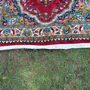 May include: A red, green and gold patterned rug with a white fringe. The rug is lying on a green grassy surface.