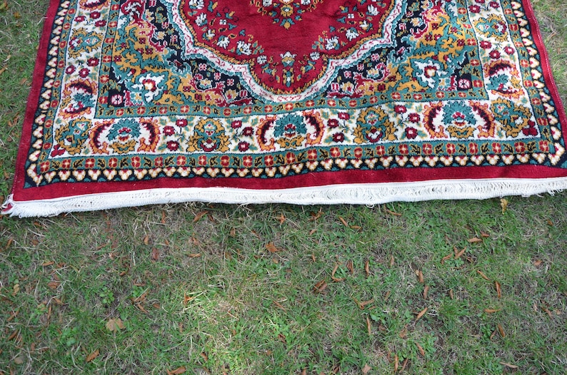 May include: A red, green and gold patterned rug with a white fringe. The rug is lying on a green grassy surface.