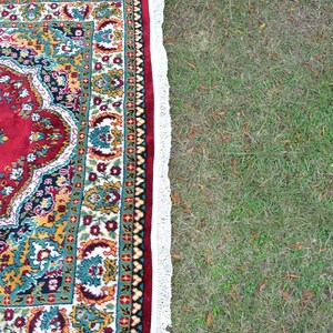 May include: A red, green and gold patterned rug with a white fringe. The rug is lying on a patch of green grass.
