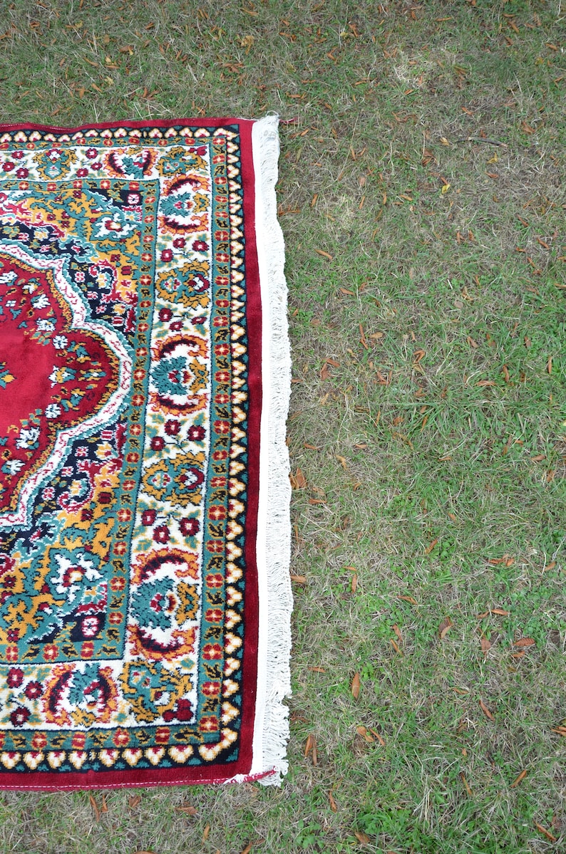 May include: A red, green and gold patterned rug with a white fringe. The rug is lying on a patch of green grass.