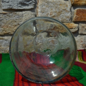 May include: A clear, round glass object with a slightly textured surface. The glass reflects the colors of the background, including red, green, and yellow. The object is set against a stone wall and a striped textile.