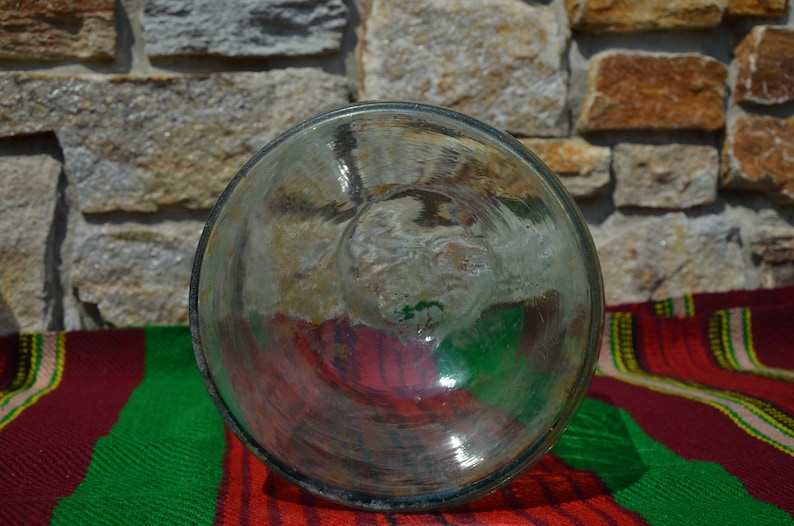 May include: A clear, round glass object with a slightly textured surface. The glass reflects the colors of the background, including red, green, and yellow. The object is set against a stone wall and a striped textile.
