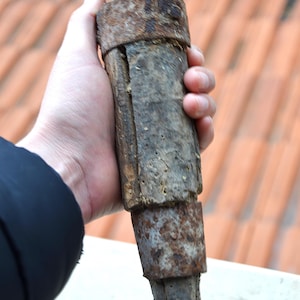 May include: A weathered, cylindrical object with a rusty, aged appearance. The object is held in a hand and appears to be made of wood and metal. The object has a tapered end and several distinct sections.