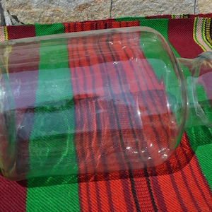 May include: A large, clear glass jar with a wide body and a narrow neck. The jar is lying on a red, green, and white striped fabric. The jar is empty and reflects the light.