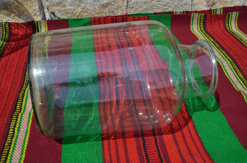 May include: A large, clear glass jar with a wide body and a narrow neck. The jar is lying on a red, green, and white striped fabric. The jar is empty and reflects the light.