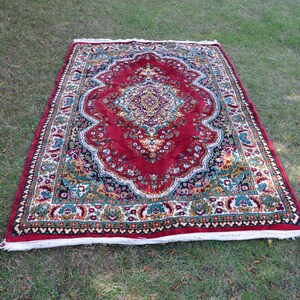 May include: A red, green and gold patterned rug with a white fringe. The rug has a floral design and is laid out on a green grassy surface.