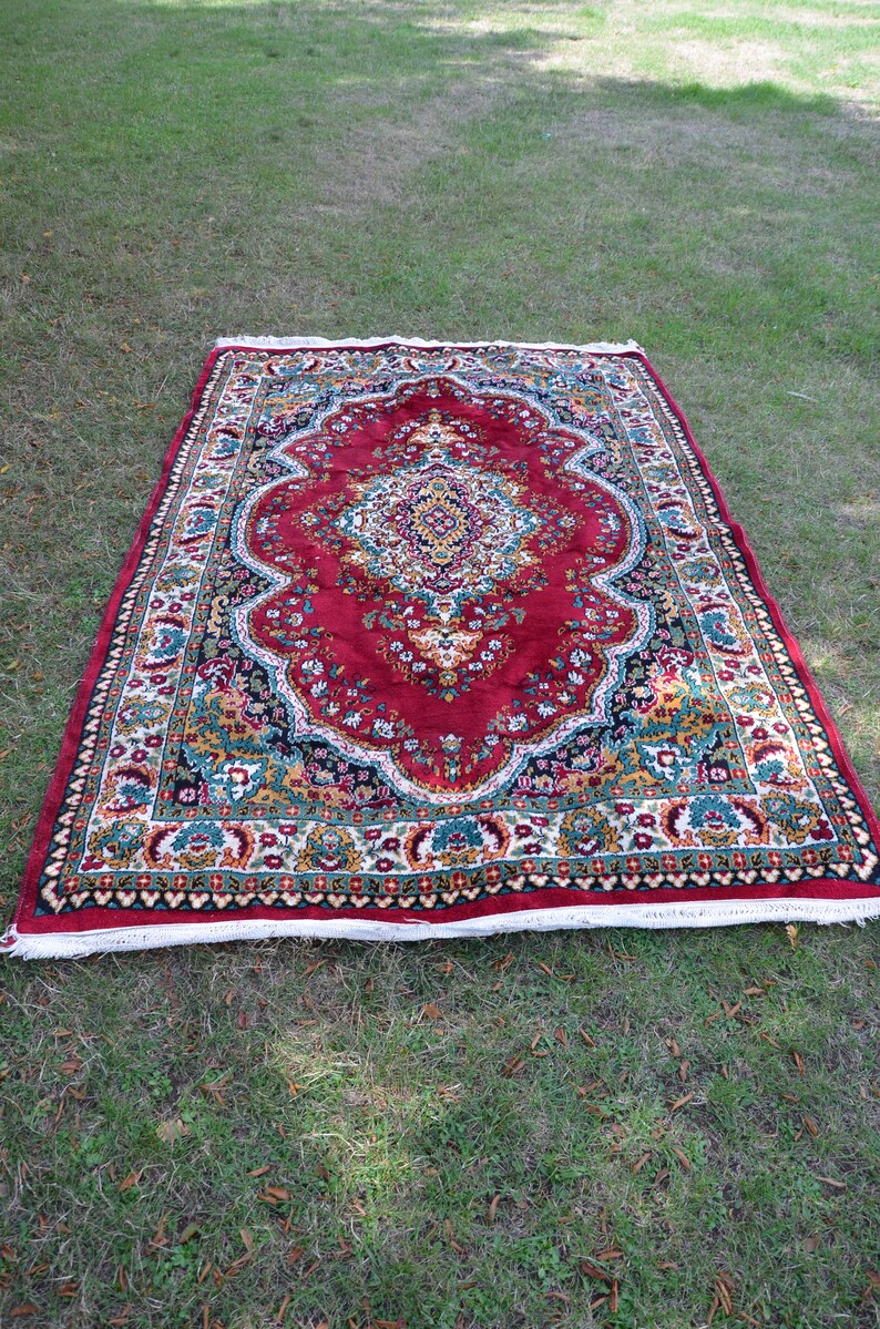 May include: A red, green and gold patterned rug with a white fringe. The rug has a floral design and is laid out on a green grassy surface.