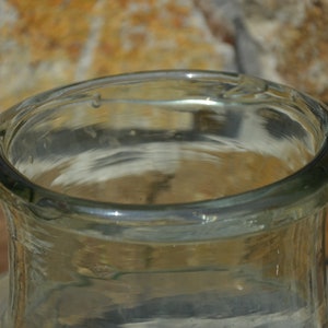 May include: Close-up of a clear glass bottle with a wide, rounded rim. The glass has a slight green tint and appears to be vintage. The bottle's opening is visible, and the background is a blurred brick wall.