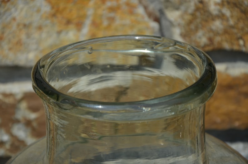 May include: Close-up of a clear glass bottle with a wide, rounded rim. The glass has a slight green tint and appears to be vintage. The bottle's opening is visible, and the background is a blurred brick wall.