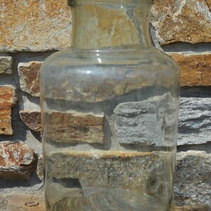 May include: A clear, cylindrical glass jar with a wide mouth and a slightly rounded base. The jar is sitting on a small, round wooden stool. The background is a stone wall with various shades of brown and gray.