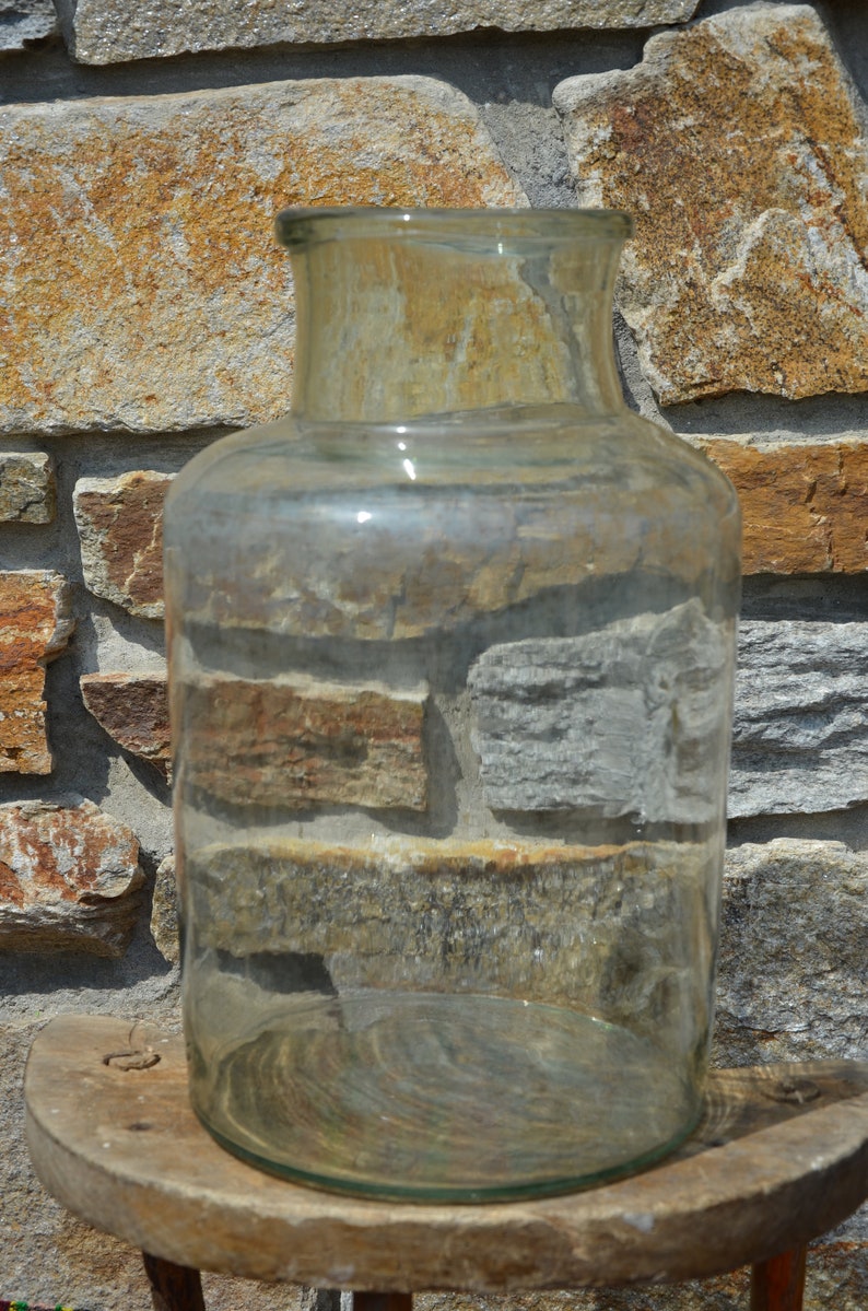 May include: A clear, cylindrical glass jar with a wide mouth and a slightly rounded base. The jar is sitting on a small, round wooden stool. The background is a stone wall with various shades of brown and gray.