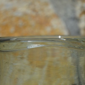 May include: Close-up of a clear glass bottle with a slightly curved rim. The bottle has a light amber tint and a smooth surface. The image highlights the bottle's shape and the play of light on the glass.