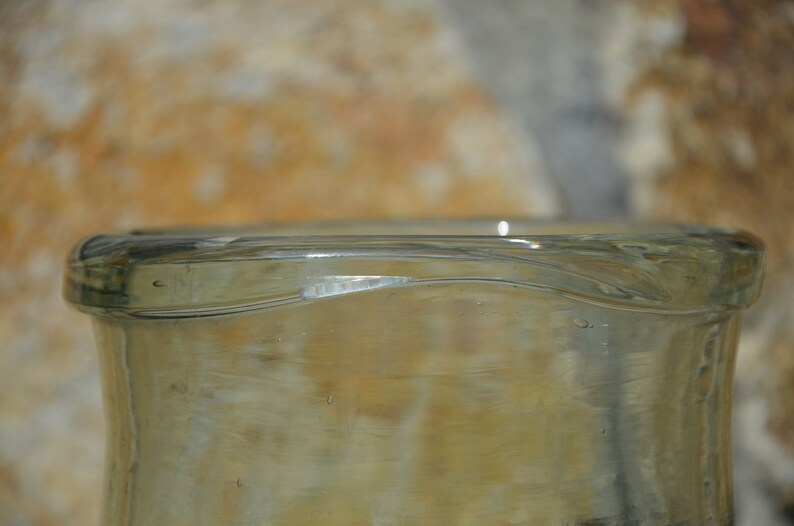 May include: Close-up of a clear glass bottle with a slightly curved rim. The bottle has a light amber tint and a smooth surface. The image highlights the bottle's shape and the play of light on the glass.