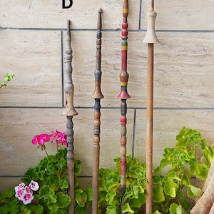 Può includere: Quattro antichi fusi di legno di vari disegni e colori. I fusi sono esposti verticalmente contro un muro strutturato, con piante in vaso in primo piano. I fusi hanno dettagli torniti decorativi e sono dipinti in tonalità di marrone, rosso, giallo e verde.