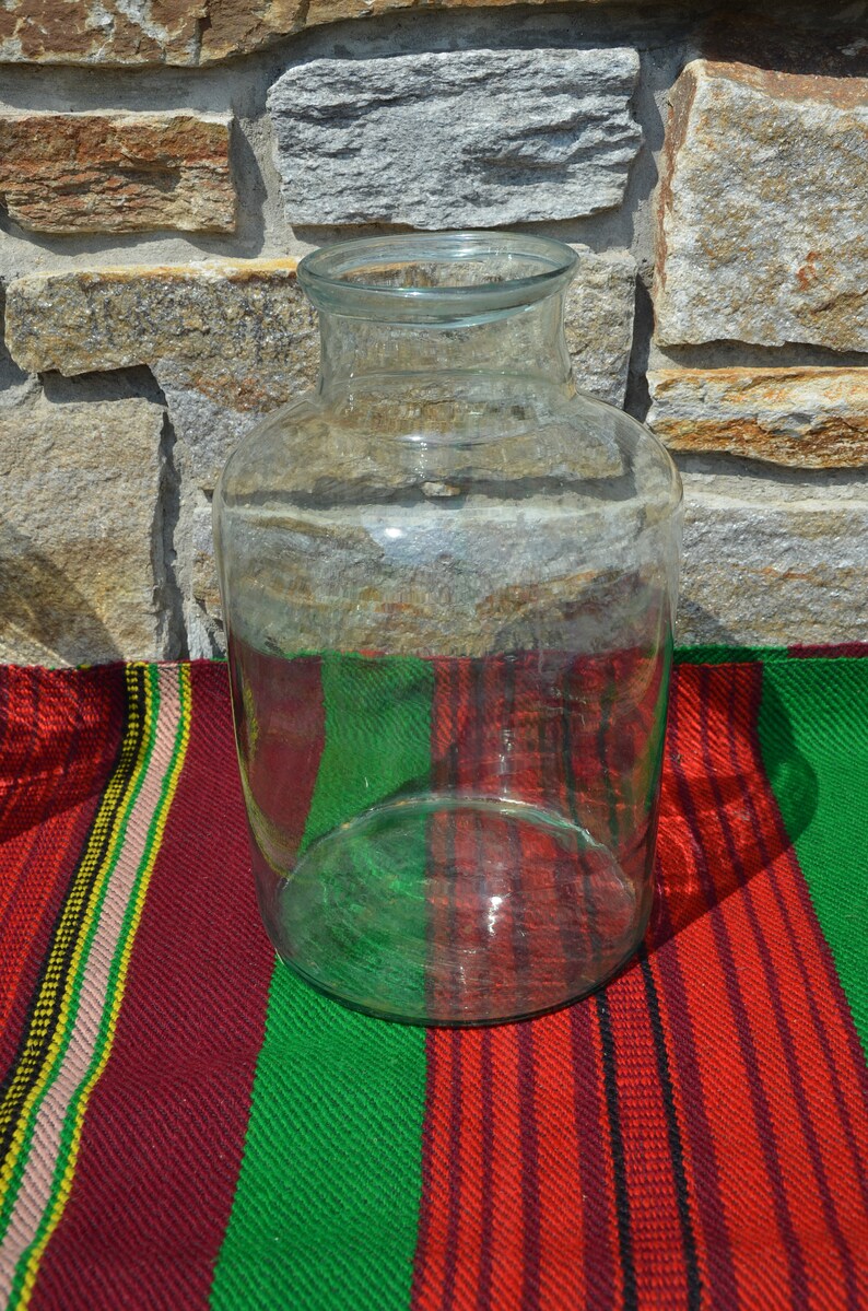 May include: A clear glass jar with a wide base and a narrow neck. The jar is sitting on a colorful striped textile with red, green, and yellow stripes. The background is a stone wall.