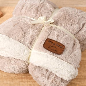 May include: A folded, plush blanket in shades of beige and white, tied with a cream ribbon. A brown leatherette patch with "MR & MRS" and "Est. 2025" is attached. The blanket has a soft, textured appearance.