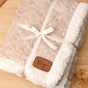 May include: A beige blanket with a textured pattern and white sherpa trim, tied with a white ribbon. A brown leather patch with the name "Bryan" is attached. The blanket is folded and ready for gifting.