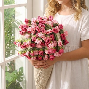 May include: A handmade crochet bouquet featuring pink and red flowers with green stems, held in a beige cone-shaped holder. The flowers are densely packed, creating a full, textured appearance. The bouquet is a unique, handcrafted item.