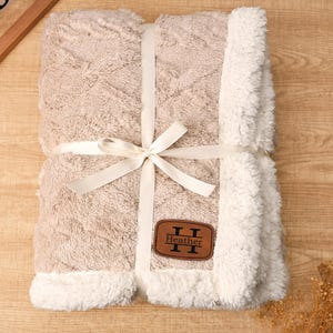 May include: A beige blanket with a white, fluffy trim and a matching ribbon tied in a bow. The blanket has a textured pattern and a brown leather patch that reads "Heather".