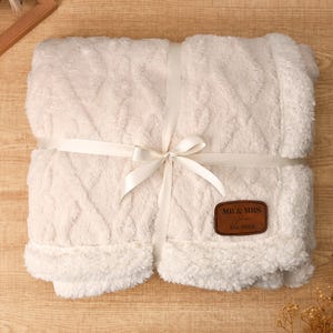 May include: Cream-colored, plush blanket with a cable knit design, wrapped with a white satin ribbon tied in a bow. A brown leather patch with "MR & MRS" and "Est. 2025" is attached. The blanket has a soft, fluffy texture.