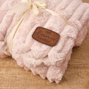 May include: A folded, pale pink plush blanket with a ribbed texture, tied with a cream ribbon and bow. A brown leather tag with the words "The Bowman Family" is attached to the blanket.