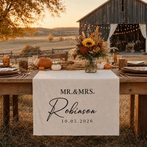 Puede incluir: Un camino de mesa de tela color crema con el texto impreso en negro MR.&MRS. Robinson y la fecha 10.05.2026. El camino se muestra sobre una mesa de madera preparada para un evento al aire libre con un centro de mesa floral y calabazas.