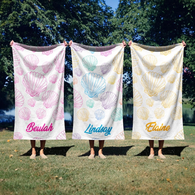 May include: Three white beach towels with seashell patterns in pink, blue, and yellow. Each towel has a name printed on it: Beulah, Lindsay, and Elaine. The towels are held up outdoors on a sunny day.