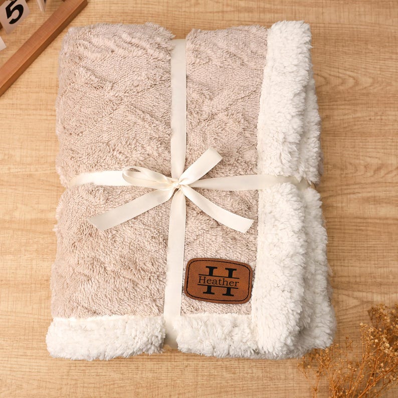 May include: A beige blanket with a white, fluffy trim and a matching ribbon tied in a bow. The blanket has a textured pattern and a brown leather patch that reads "Heather".