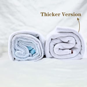 May include: Two rolled white towels, one with a blue interior, are displayed on a white surface. The text "Thicker Version" is above the right towel, indicating a comparison of thicknesses. The towels are neatly rolled, showcasing their texture.