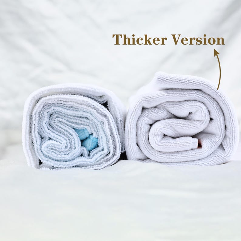 May include: Two rolled white towels, one with a blue interior, are displayed on a white surface. The text "Thicker Version" is above the right towel, indicating a comparison of thicknesses. The towels are neatly rolled, showcasing their texture.
