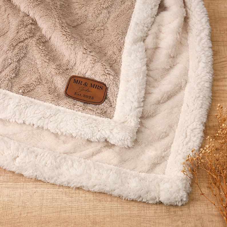 May include: A plush, beige blanket with a cream-colored, fluffy border. The blanket features a textured pattern and a brown leather patch that reads "MR & MRS, Est. 2025". The blanket is folded, showcasing its soft texture.