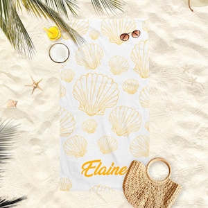 May include: White beach towel with a pattern of gold seashells. The name "Elaine" is printed in gold script. Sunglasses and a straw bag are next to the towel on the sand.