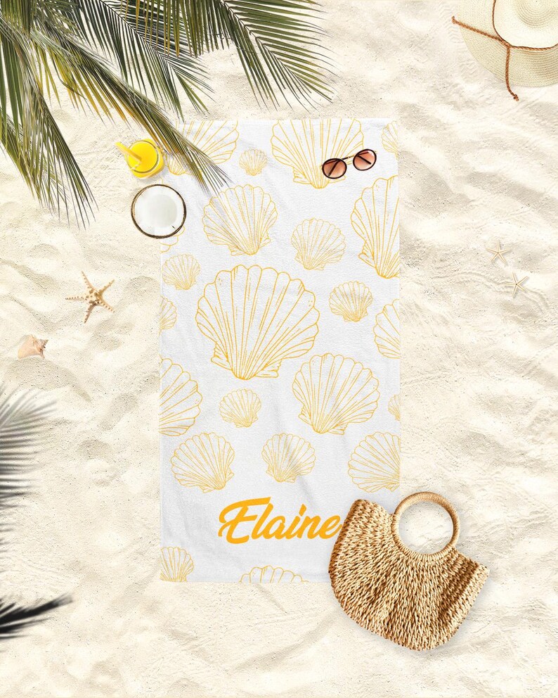 May include: White beach towel with a pattern of gold seashells. The name "Elaine" is printed in gold script. Sunglasses and a straw bag are next to the towel on the sand.