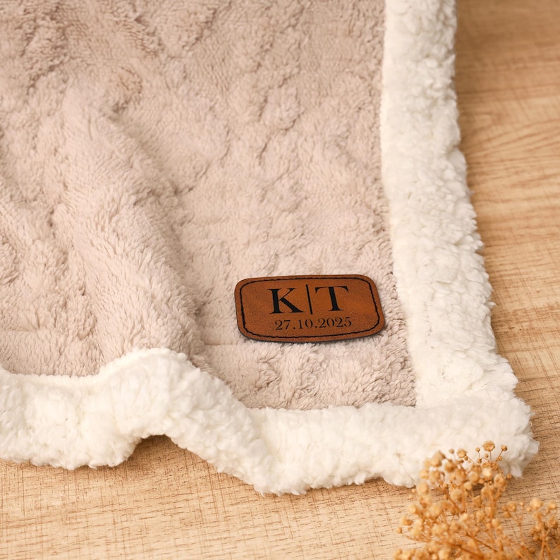 May include: A beige, plush blanket with a textured pattern and a white, fluffy border. A brown leather patch with the initials "KT" and the date "27.10.2025" is sewn on the blanket. The blanket is folded, showing a corner.