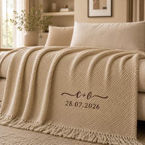 May include: A beige textured knit throw blanket with fringe edges, featuring dark brown embroidered text that reads C + O and the date 28.07.2026. The blanket has a consistent popcorn-like stitch pattern throughout its surface.