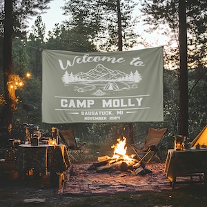 May include: A green camping banner with white text that reads "Welcome to Camp Molly, Saugatuck, MI, November 2024". The banner is hanging between two trees in a campsite. There is a campfire in the foreground with two chairs and a table.