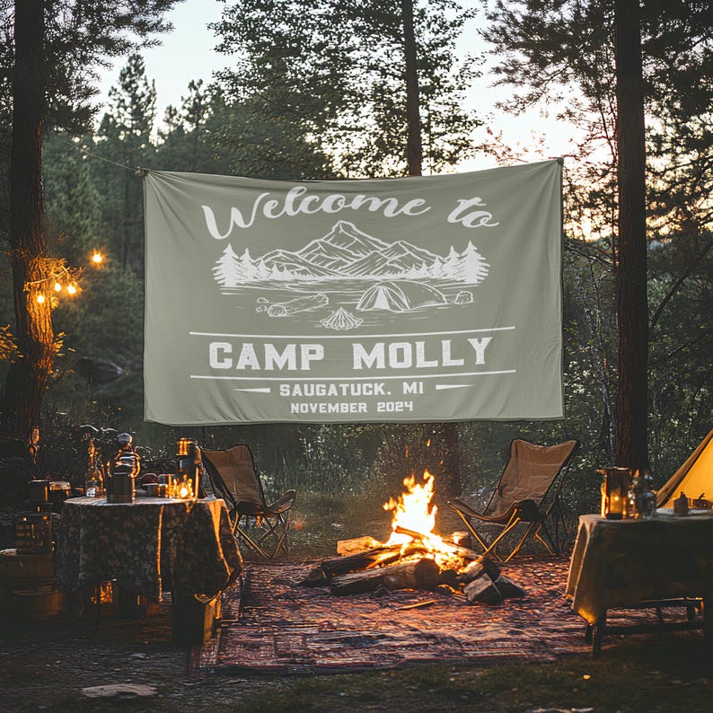 May include: A green camping banner with white text that reads "Welcome to Camp Molly, Saugatuck, MI, November 2024". The banner is hanging between two trees in a campsite. There is a campfire in the foreground with two chairs and a table.