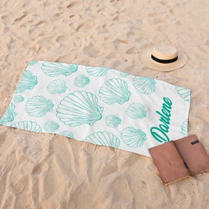 May include: White beach towel with a teal seashell pattern and the name "Darlene" in teal script. A straw hat with a black band and two brown notebooks are on the sand.