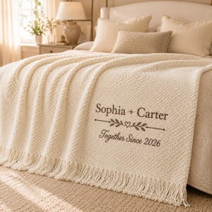 Pode incluir: Uma manta texturizada de cor creme com franjas, apresentando um texto bordado em castanho escuro que diz Sophia + Carter, um divisor decorativo de folhas e coração, e a frase Together Since 2026, disposta sobre uma cama.