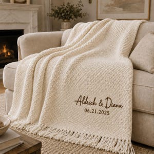 May include: A cream-colored, textured knit throw blanket featuring fringe trim along the bottom edge. The blanket is embroidered with the brown text "Aldrich & Diana" followed by the date "06.21.2025" in a script and sans-serif font.